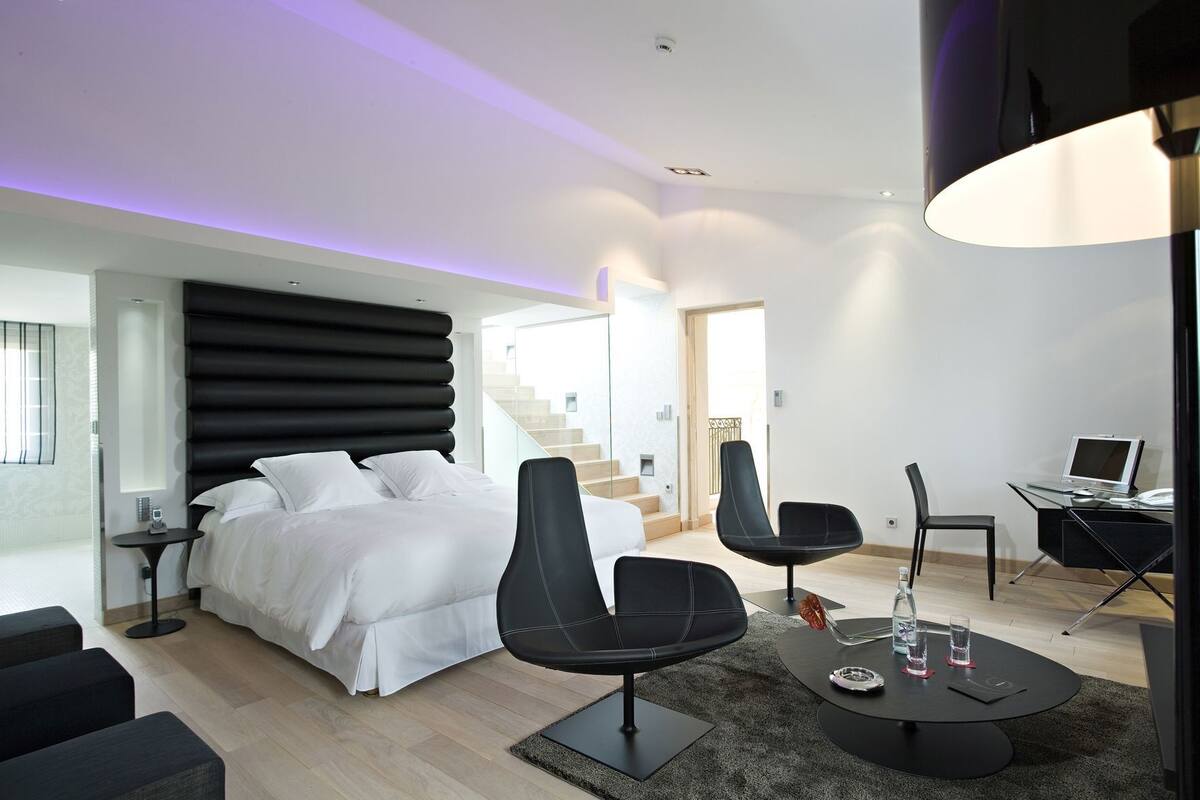 Een moderne hotelkamer met een bed, twee zwarte stoelen, een bureau met een laptop en een trap naar een verdieping erboven.