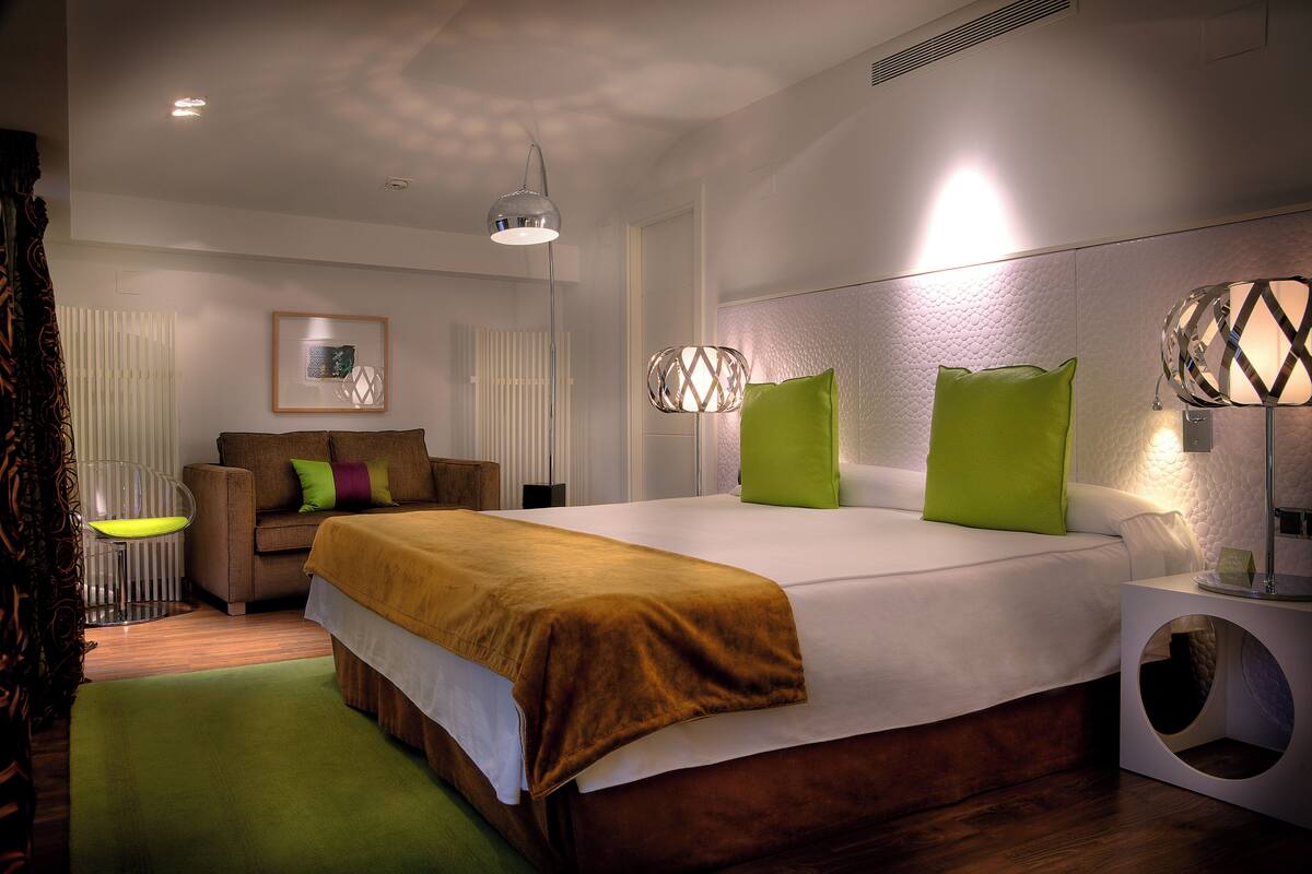 A modern hotel room with a large bed, a sofa, a chair, and a unique circular side table.