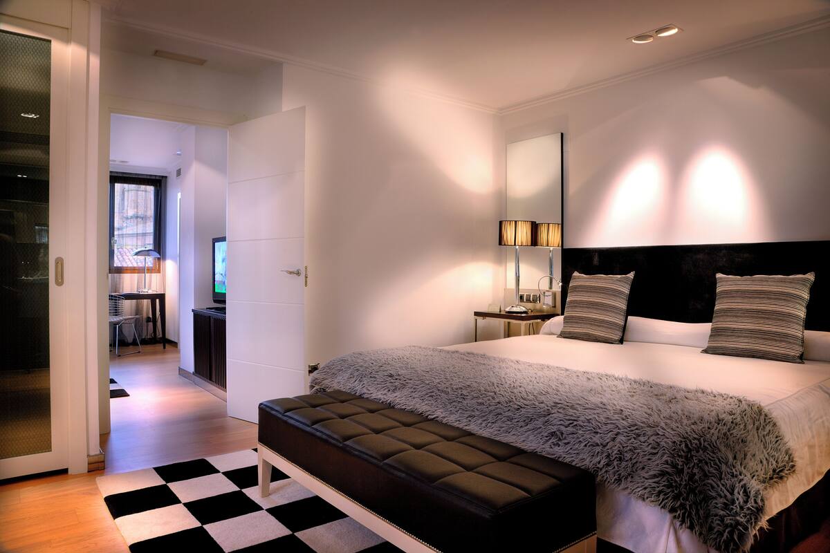 A modern bedroom with a large bed, a bench, a desk, and a TV.