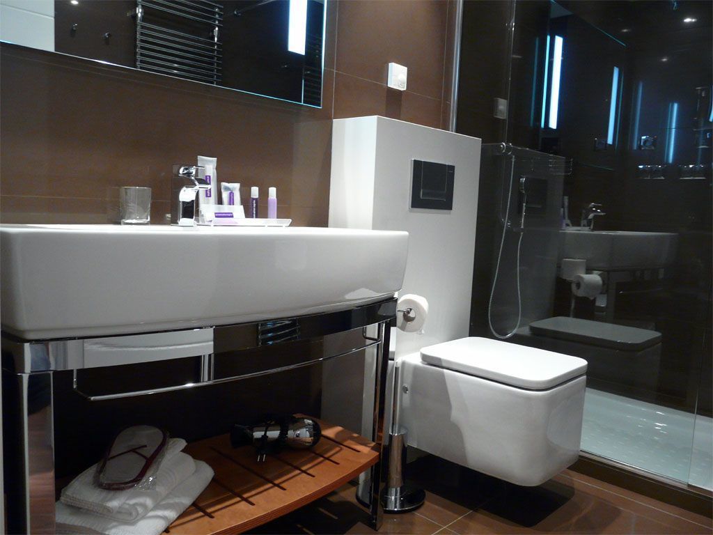 Een moderne badkamer met een wit wastafel, spiegel en toilet. Er is een glazen douchecabine en een klein plankje met toiletartikelen.