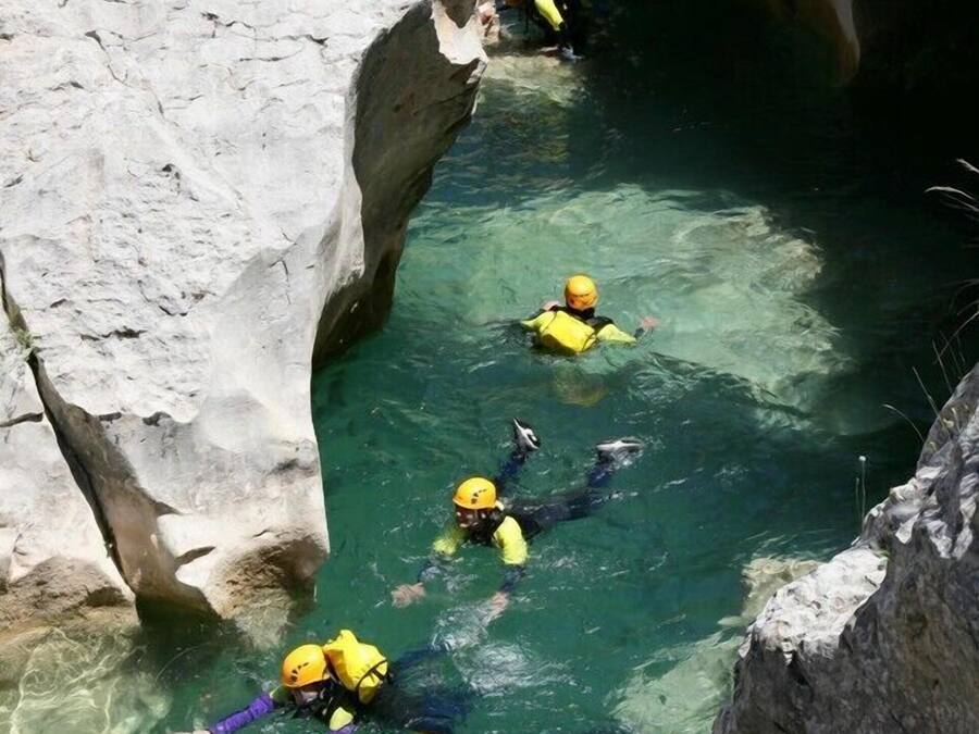 A group of people in yellow helmets and life vests swimming in a narrow, rocky waterway.