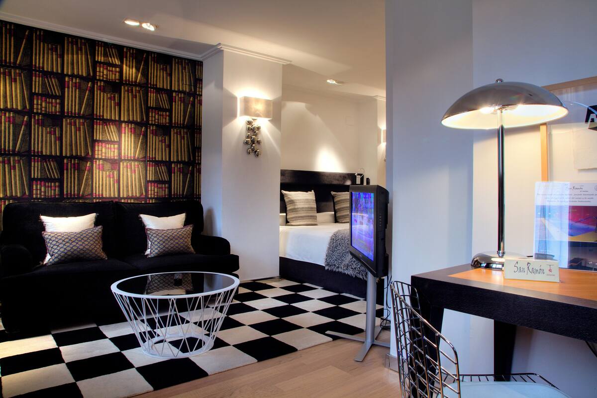 A modern hotel room with a black and white checkered floor, a black sofa with patterned cushions, a round glass coffee table, a flat-screen TV, a desk with a lamp, and a bookshelf filled with books.