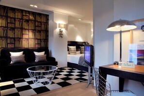 A modern hotel room with a black and white checkered floor, a black sofa with patterned cushions, a round glass coffee table, a flat-screen TV, a desk with a lamp, and a bookshelf filled with books.