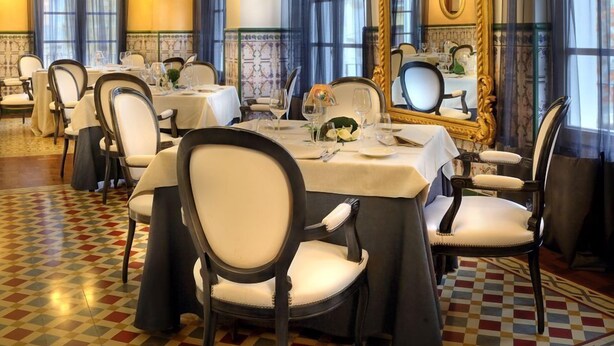 Un comedor formal con mesas dispuestas para el servicio, un espejo grande y ornamentado, y un piso con diseño.