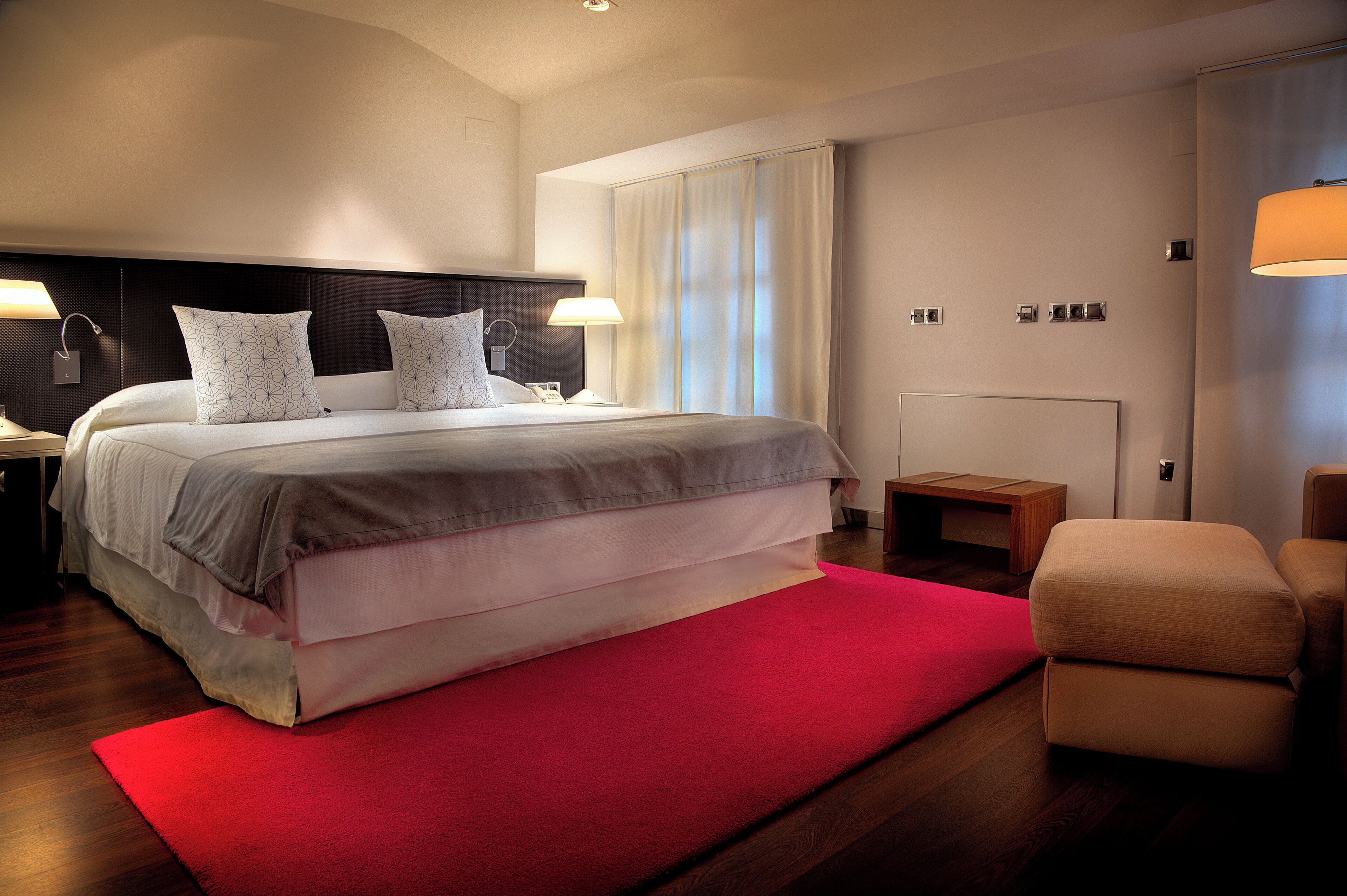 Una habitación de hotel moderna con una cama grande, lámparas de noche, un tapete rojo y una mesita de noche de madera.
