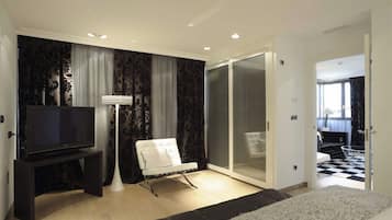 A modern bedroom with a flat-screen TV, a white chair, a large window with curtains, and a sliding glass door leading to a balcony.