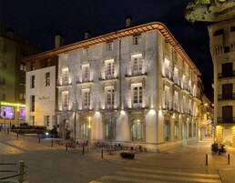 Un edificio histórico con varios balcones y ventanas amplias, rodeado de una plaza pavimentada con mesas al aire libre.