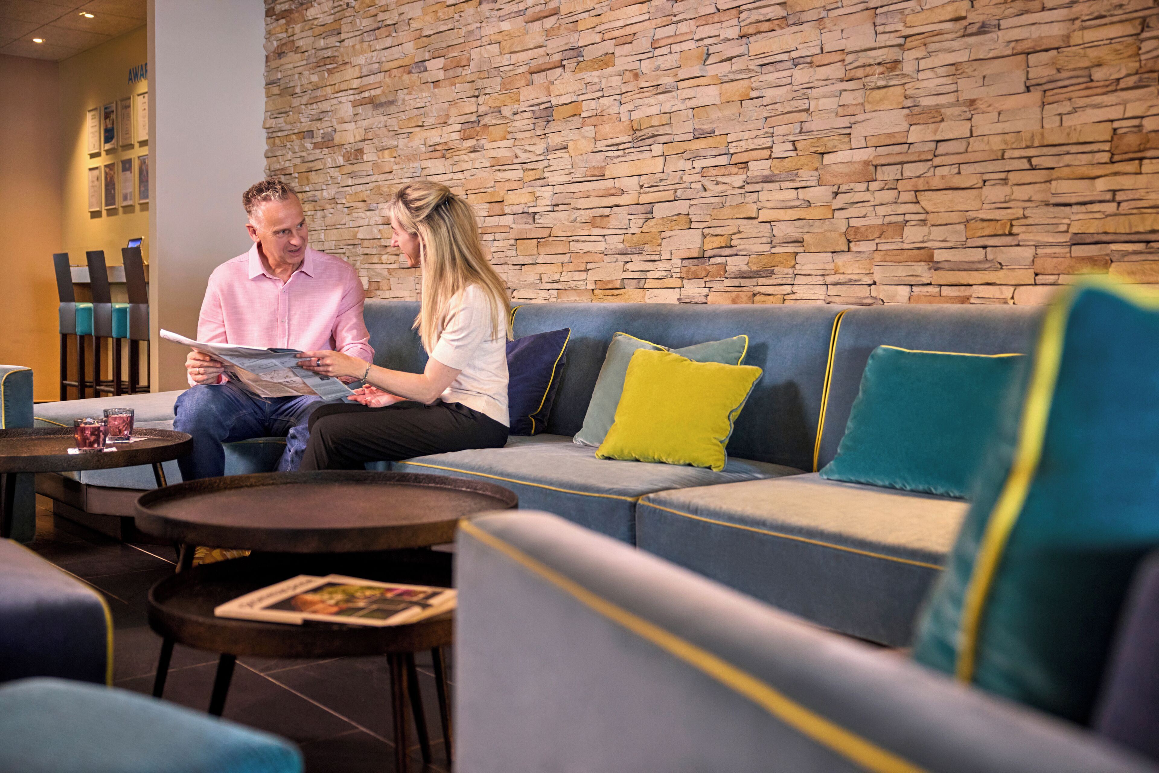 A modern lounge area with a stone wall, a blue sofa with yellow and teal cushions, and a small table with drinks.