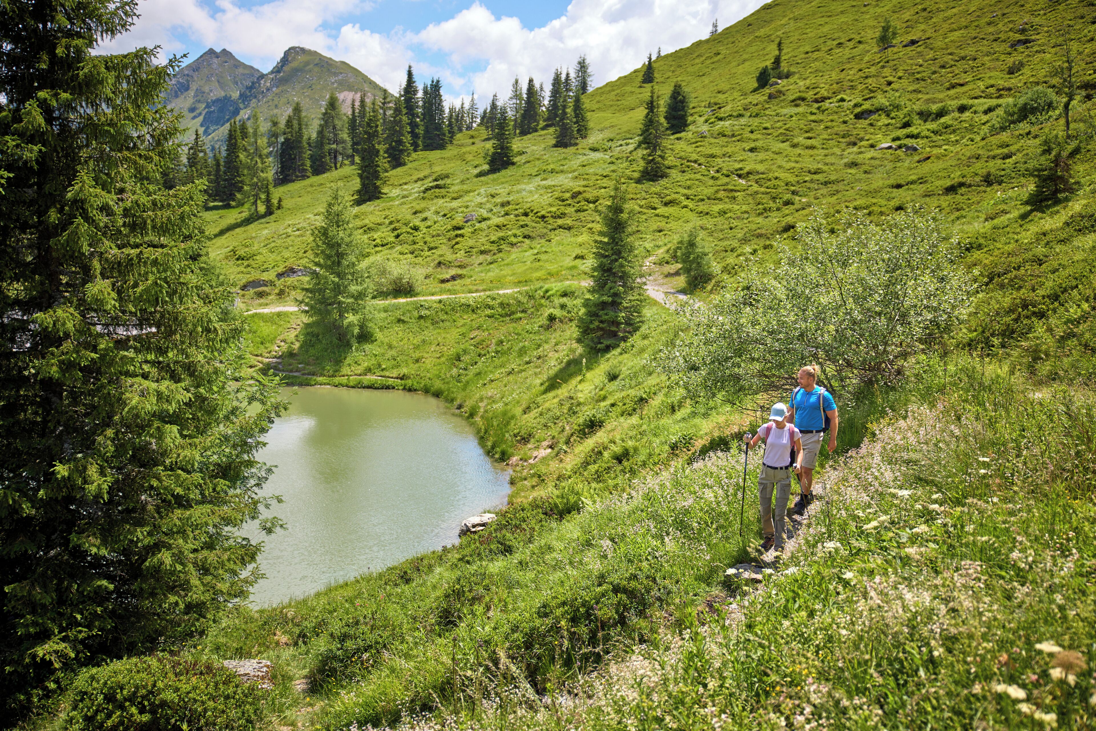 A mountainous landscape with a lake, a hiking trail, and two people walking.