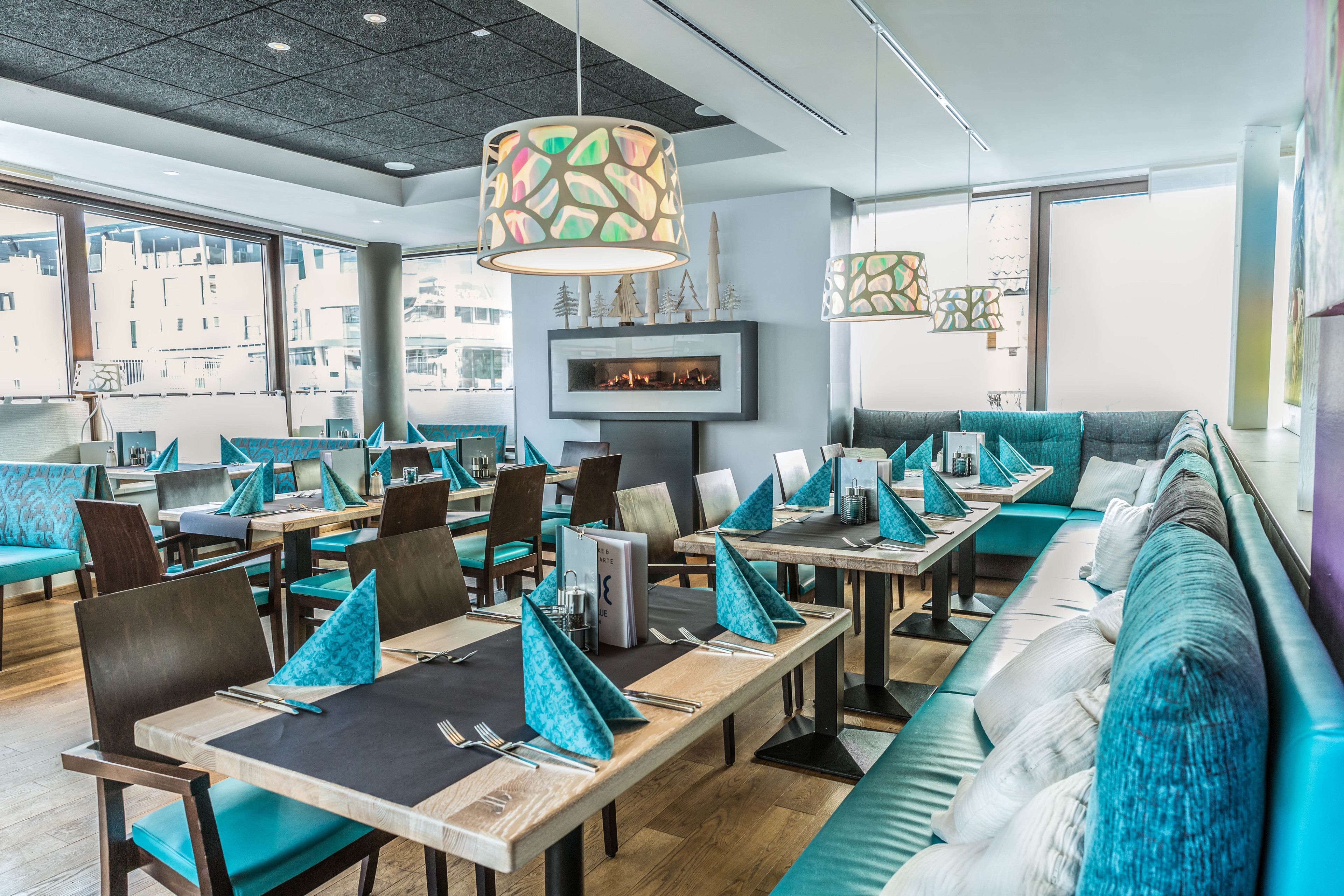 Un restaurant moderne avec des sièges aux couleurs turquoise et gris, une cheminée et de grandes fenêtres offrant une vue sur la ville.
