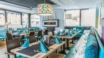 Un restaurant moderne avec des sièges aux couleurs turquoise et gris, une cheminée et de grandes fenêtres offrant une vue sur la ville.