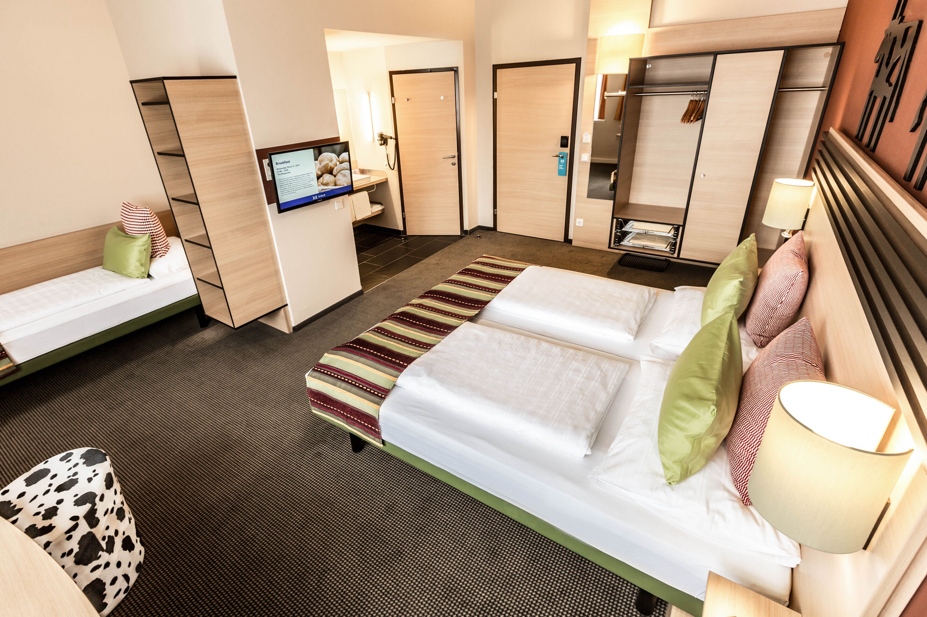 A hotel room with a large bed, a smaller bed, a TV, a wardrobe, and a desk with a lamp.