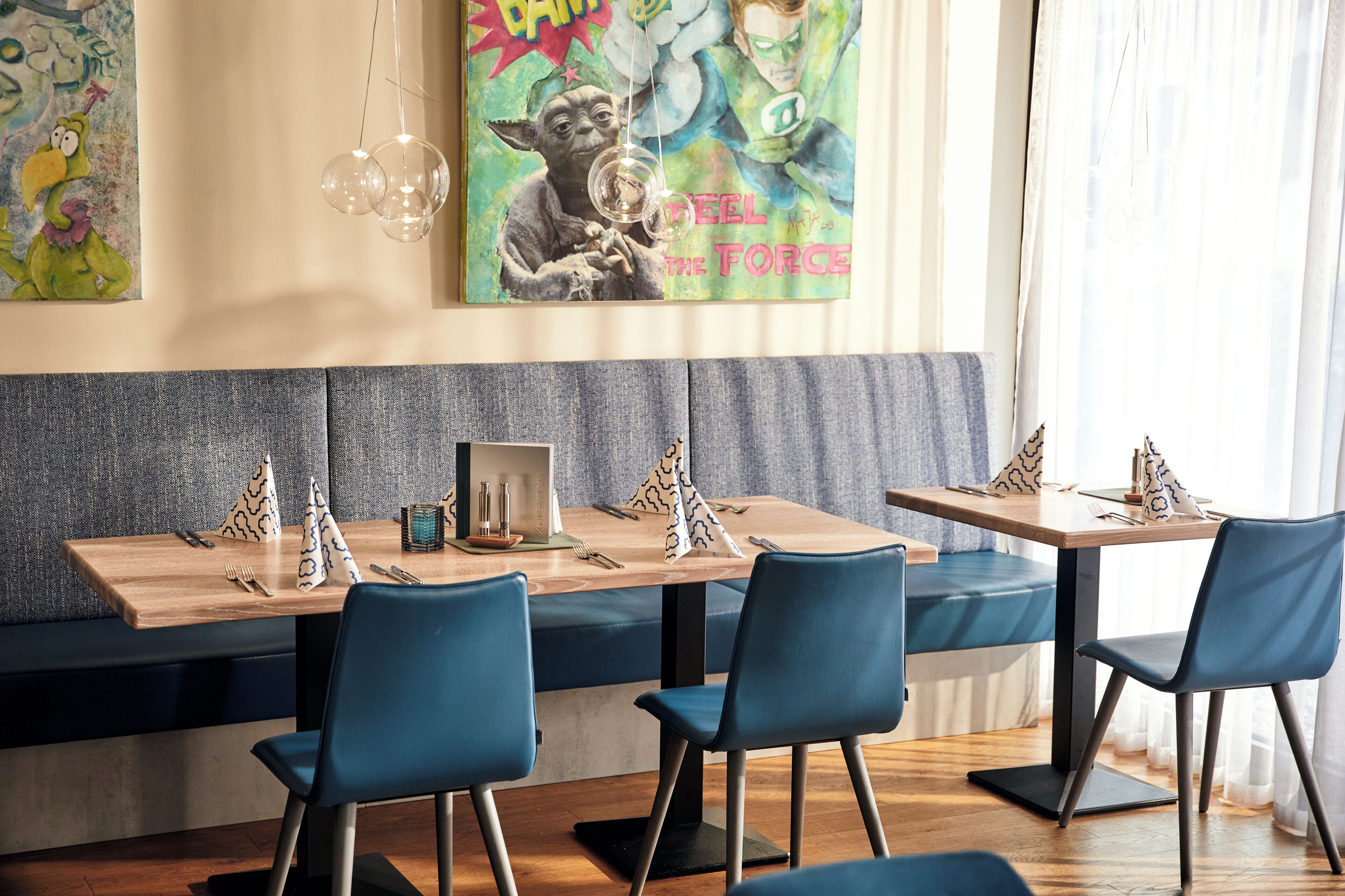 Interior de restaurante con mesas de madera, sillas azules y una pared con arte colorido.