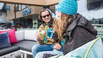 Ein Paar sitzt auf einer grauen Outdoor-Sofa mit Kissen und hält Getränke mit Limette und Eis. Im Hintergrund ist ein Hotelschild sichtbar.