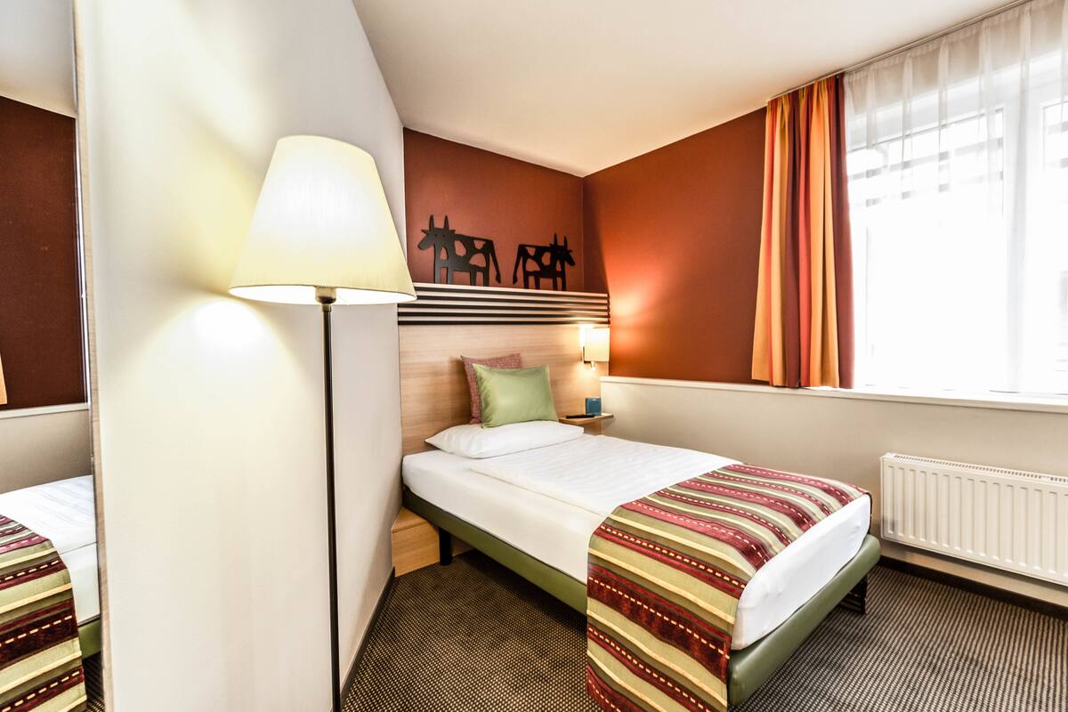 Een hotelkamer met een bed, nachtkastje, lamp, raam met gordijnen en een wand met silhouet van een koe.
