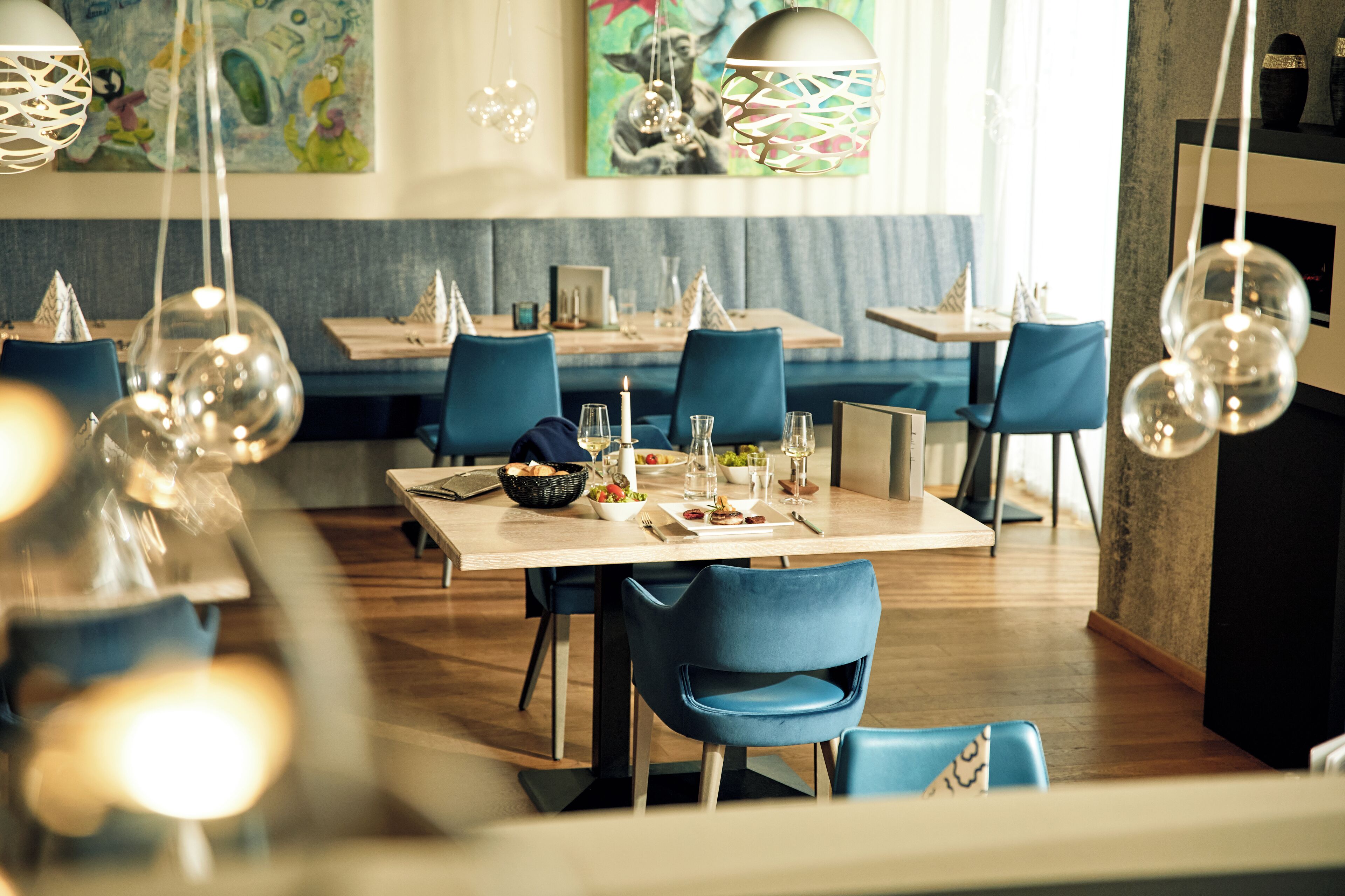 A dining area with tables set for two, blue chairs, hanging spherical light fixtures, and a painting on the wall.