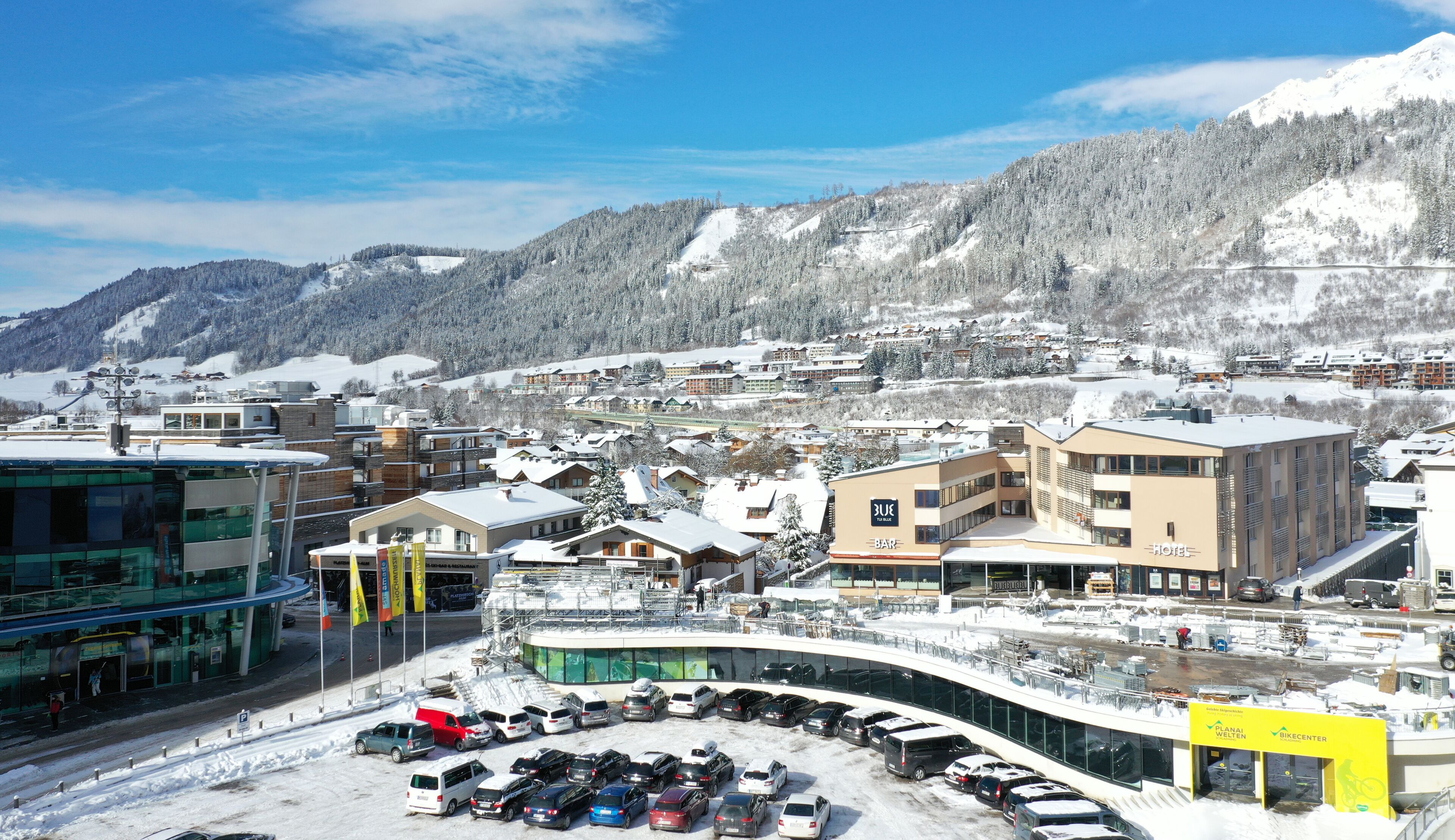 A ski resort town with a mix of modern and traditional buildings, a parking area, and a mountain backdrop.