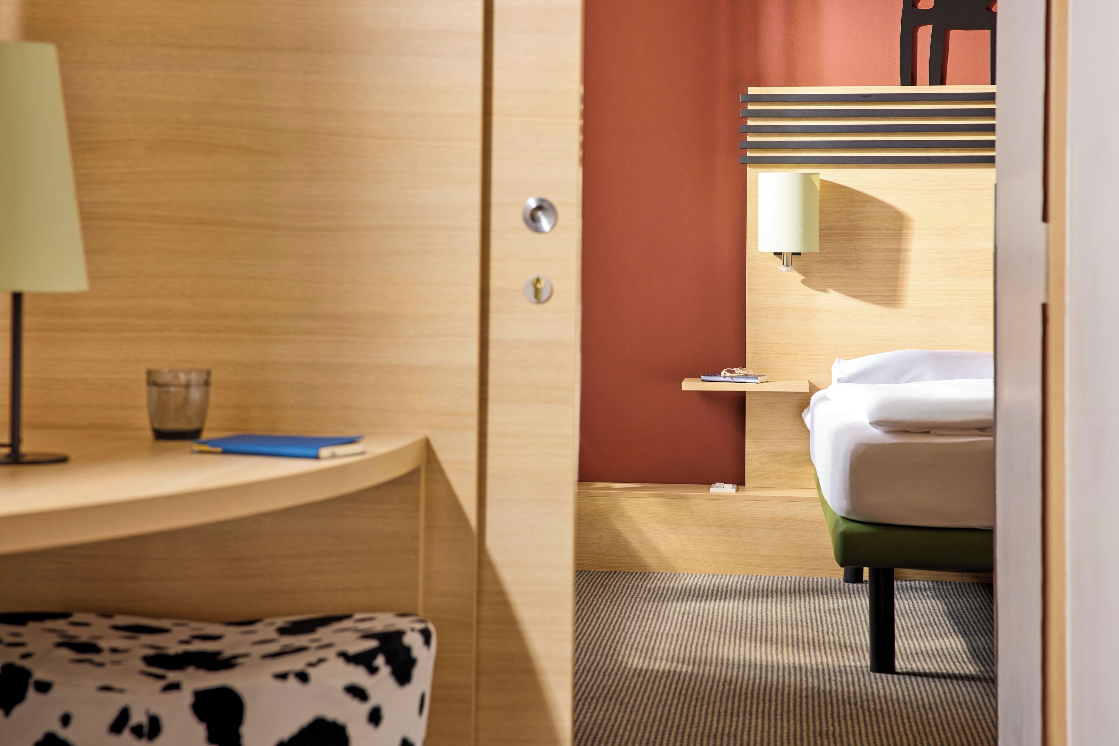 Ein Hotelzimmer mit einem Holztisch, einer Lampe, einem Glas, einem blauen Notizbuch, einem Bett mit weißer Bettwäsche und einem grünen Sessel.
