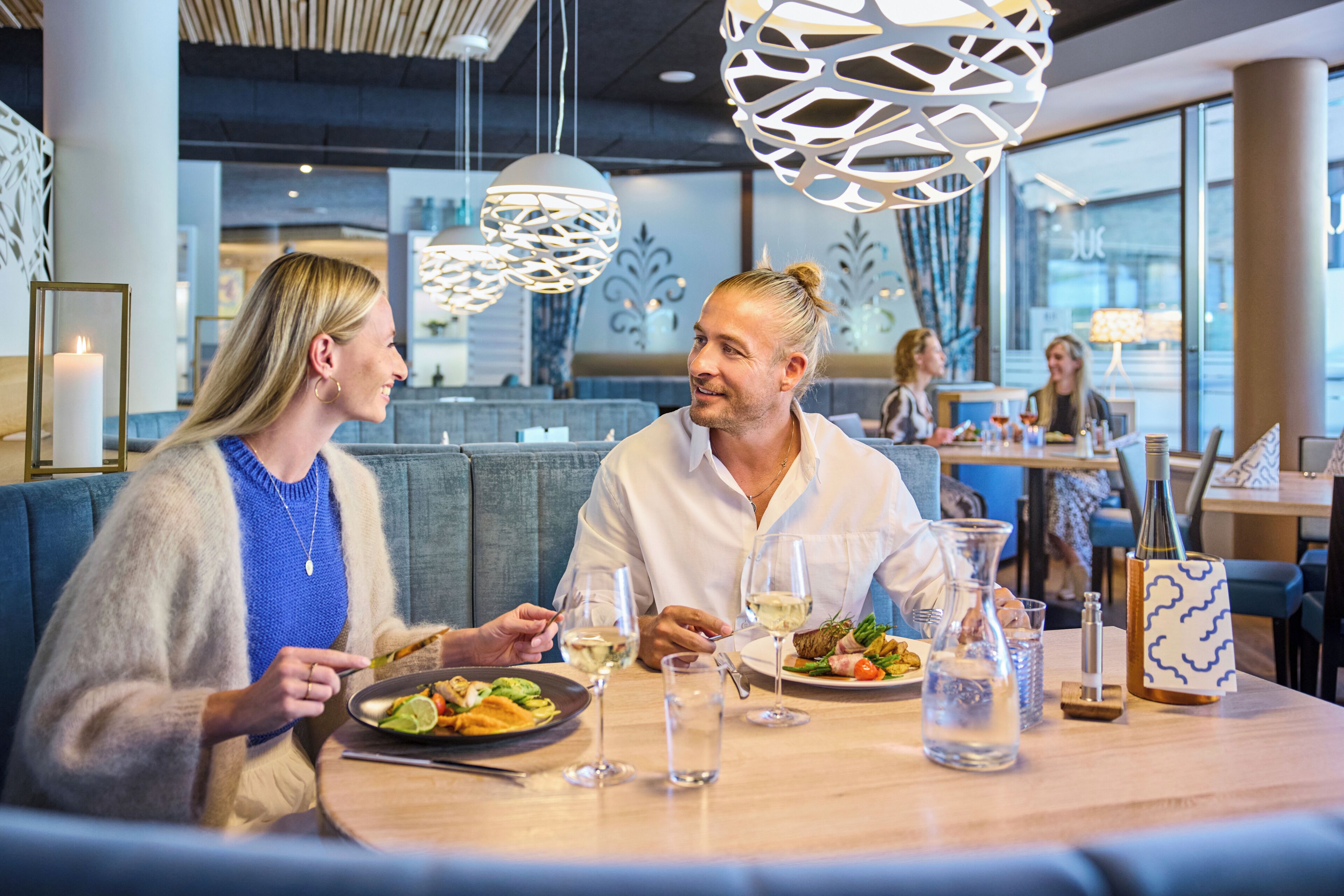 Un ambiente de restaurante con una mesa preparada para dos personas, que incluye una comida, copas de vino y una jarra de agua.