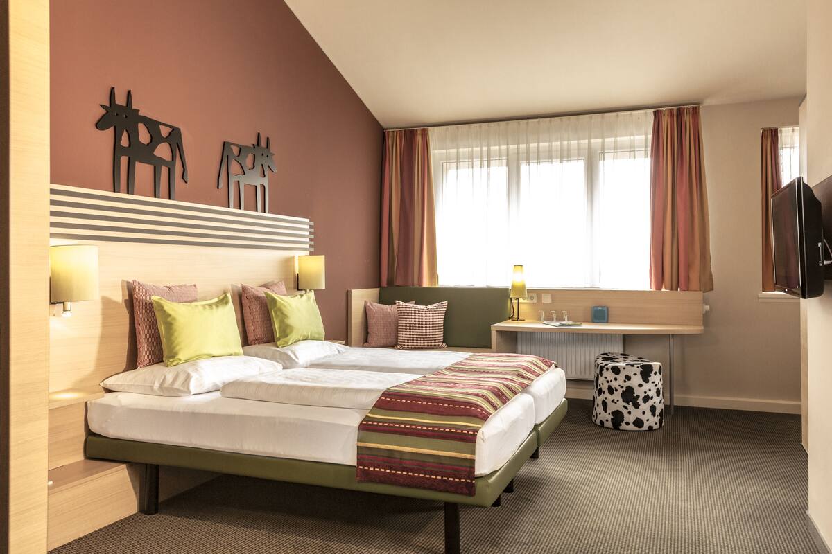 Een tweepersoonsbed met een gestreept dekbed, een nachtkastje met een lamp en een aan de muur bevestigde televisie.
