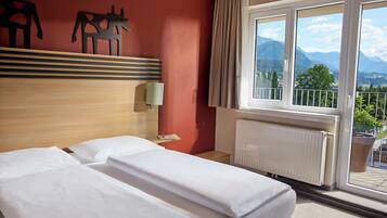 A hotel room with a bed, a nightstand, a lamp, a window with a view of mountains, and a radiator.