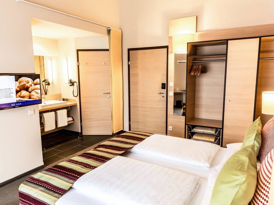 A hotel room with a bed, a bathroom with a mirror, a sink, and a TV displaying breakfast information.