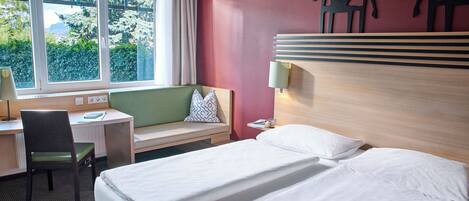 A hotel room with a bed, a desk, a chair, a window with a view of greenery, and a wall with a cutout of a giraffe.