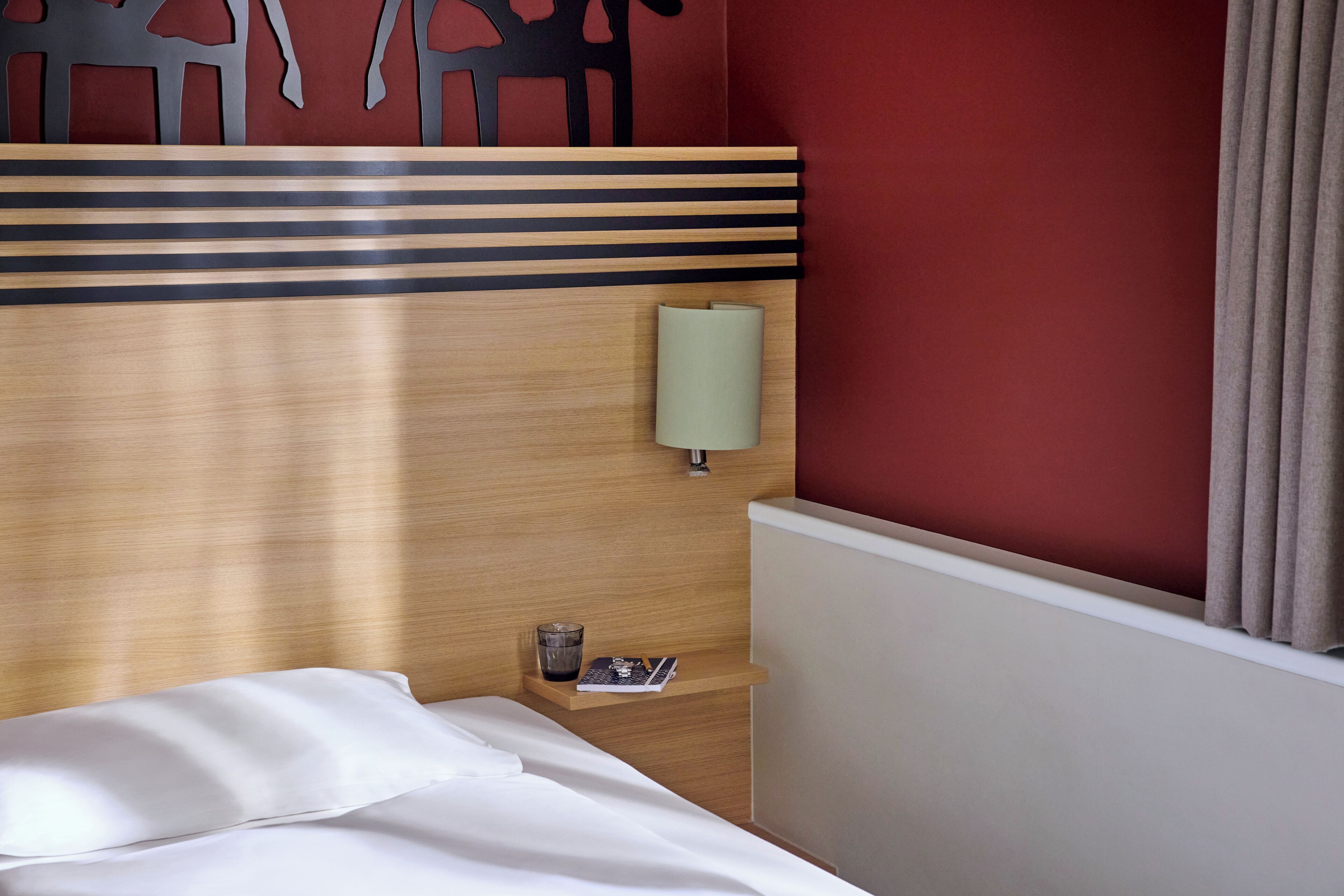 Ein Hotelzimmer mit einem hölzernen Kopfteil, einem Nachttisch mit Lampe, einem Glas Wasser und einer Zeitschrift.