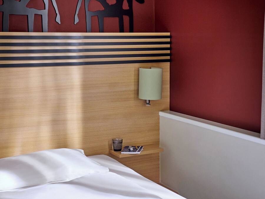 Habitación de hotel con cabecera de madera, mesita de noche con lámpara, vaso de agua y una revista.