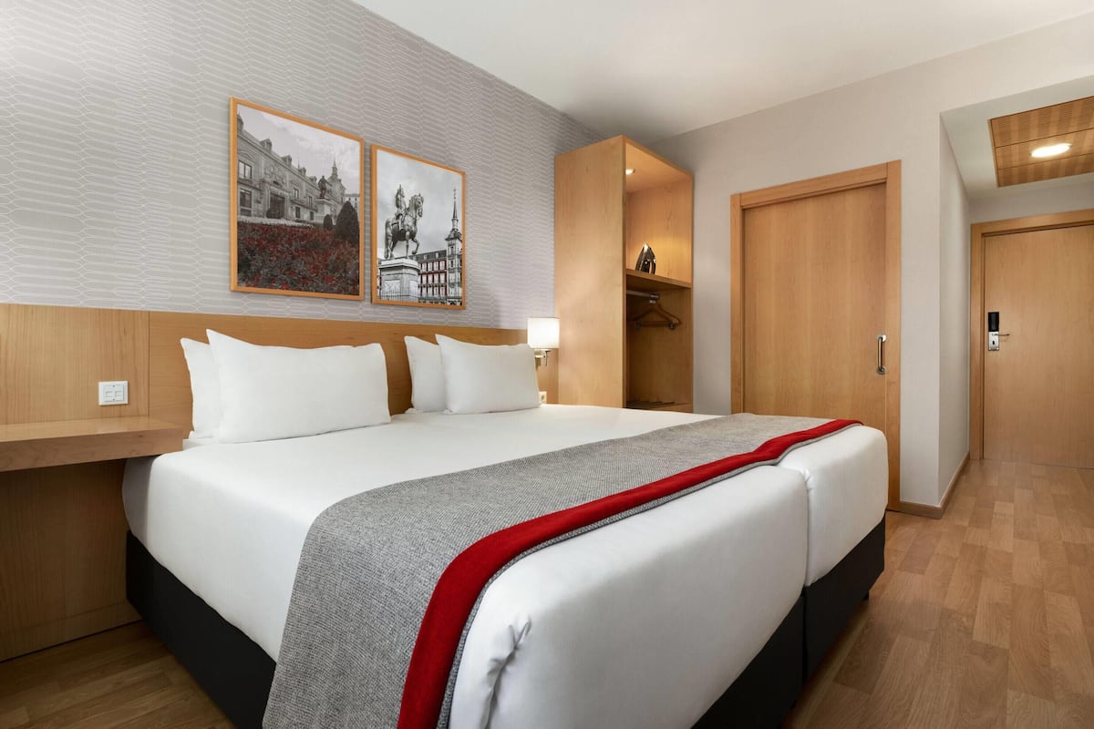 A hotel room with a large bed, a wooden headboard, a nightstand, a wall-mounted lamp, a wardrobe, and two framed pictures on the wall.