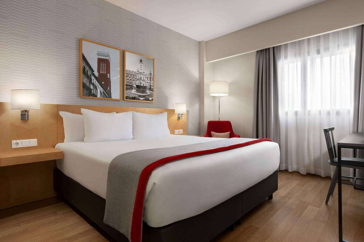 A hotel room with a large bed, two bedside tables with lamps, a desk with a chair, and a large window with curtains.