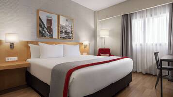 A hotel room with a large bed, two bedside tables, a desk, and a chair. There are two wall-mounted lamps and a framed picture above the bed.