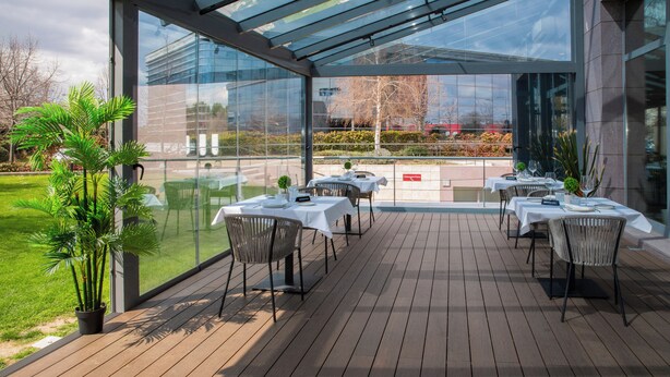 Un área de comedor al aire libre con mesas y sillas, un cerramiento de cristal y vistas del paisaje circundante.