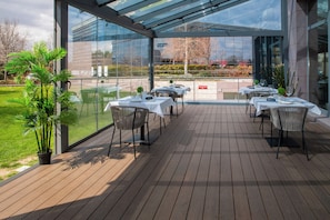 Un área de comedor al aire libre con mesas y sillas, un cerramiento de cristal y vistas del paisaje circundante.