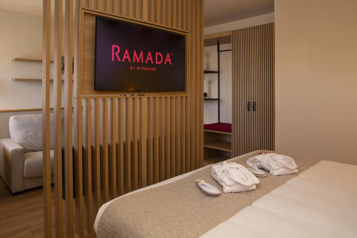 A hotel room with a bed, a TV mounted on a wooden panel, a sofa, and a wardrobe.