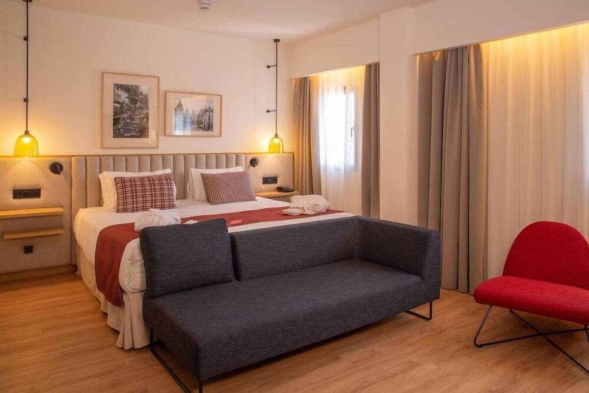 A hotel room with a bed, a grey sofa, a red chair, and two wall-mounted lamps.