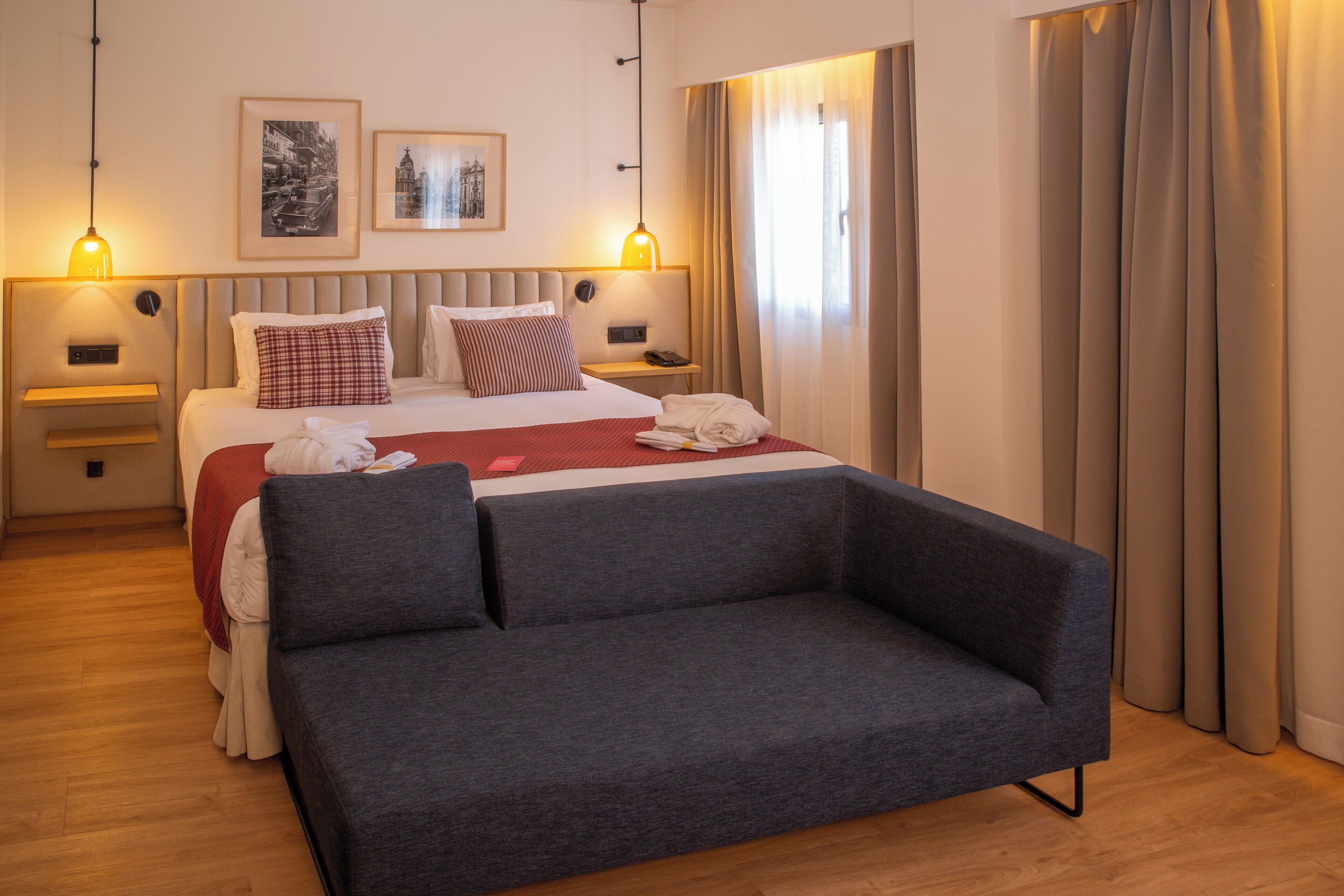 Un hotel moderno con una cama grande, un sofá gris, dos mesitas de noche con lámparas y dos cuadros en la pared.