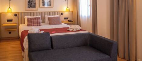 Un hotel moderno con una cama grande, un sofá gris, dos mesitas de noche con lámparas y dos cuadros en la pared.