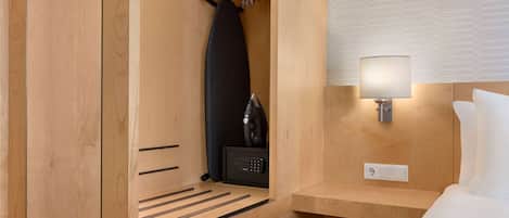 Una habitación de hotel con un armario de madera, una tabla de planchar negra, una caja fuerte negra y una lámpara de noche.