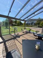 A covered outdoor patio with a glass roof, furnished with tables and chairs, and a view of a grassy area and buildings in the background.