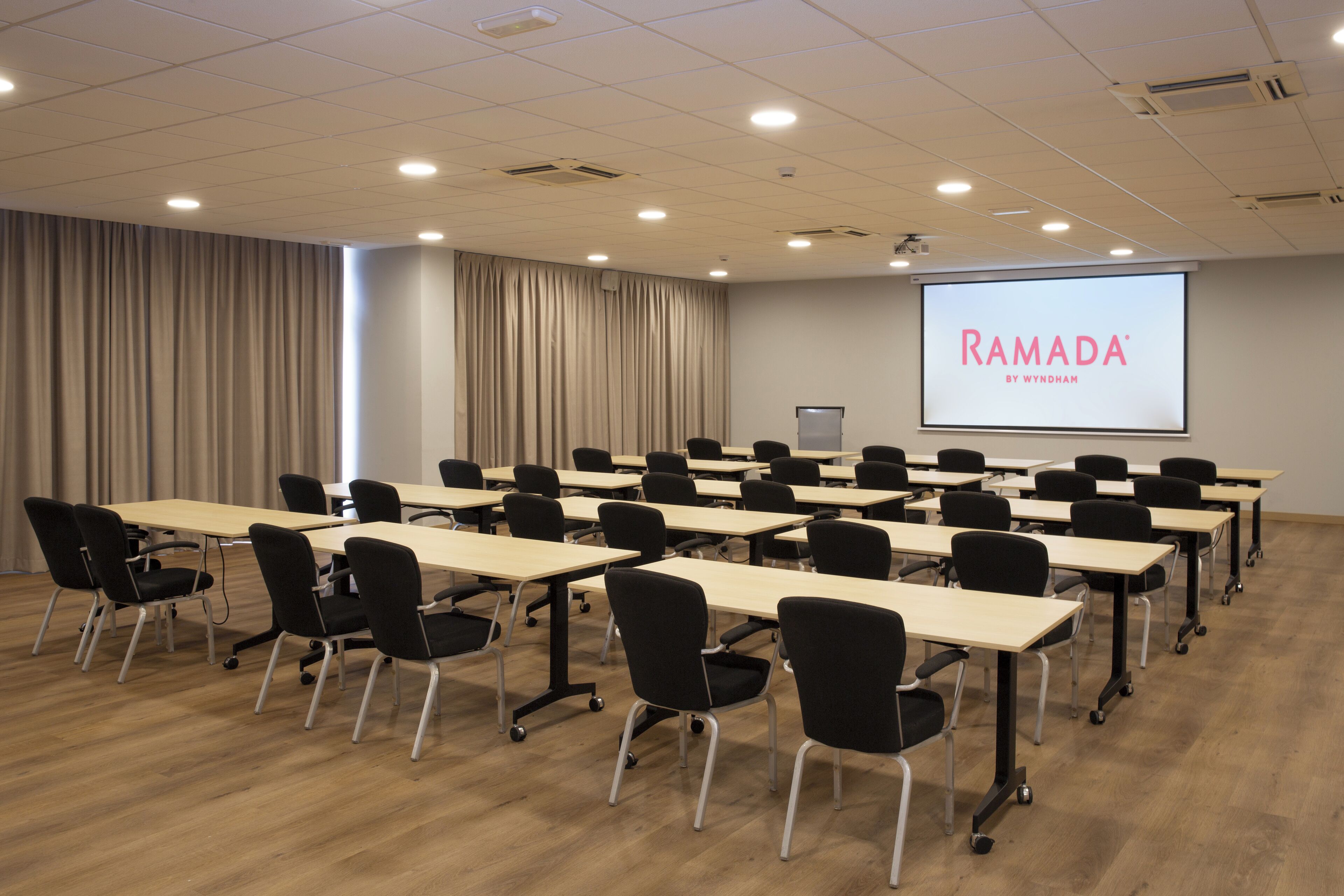 Una sala de conferencias con varias mesas y sillas, una pantalla de proyector mostrando "RAMADA BY WYNDHAM" y una cortina en la pared izquierda.