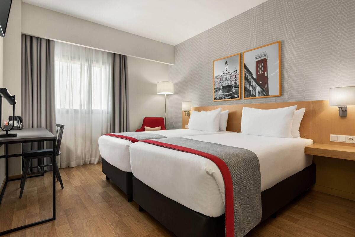 A hotel room with two beds, a desk, and a chair. There are two wall-mounted lamps and a window with curtains.