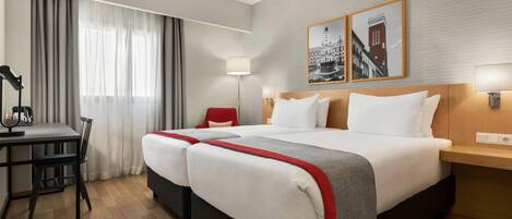 A hotel room with two beds, a desk, and a chair. There are two wall-mounted lamps and a window with curtains.