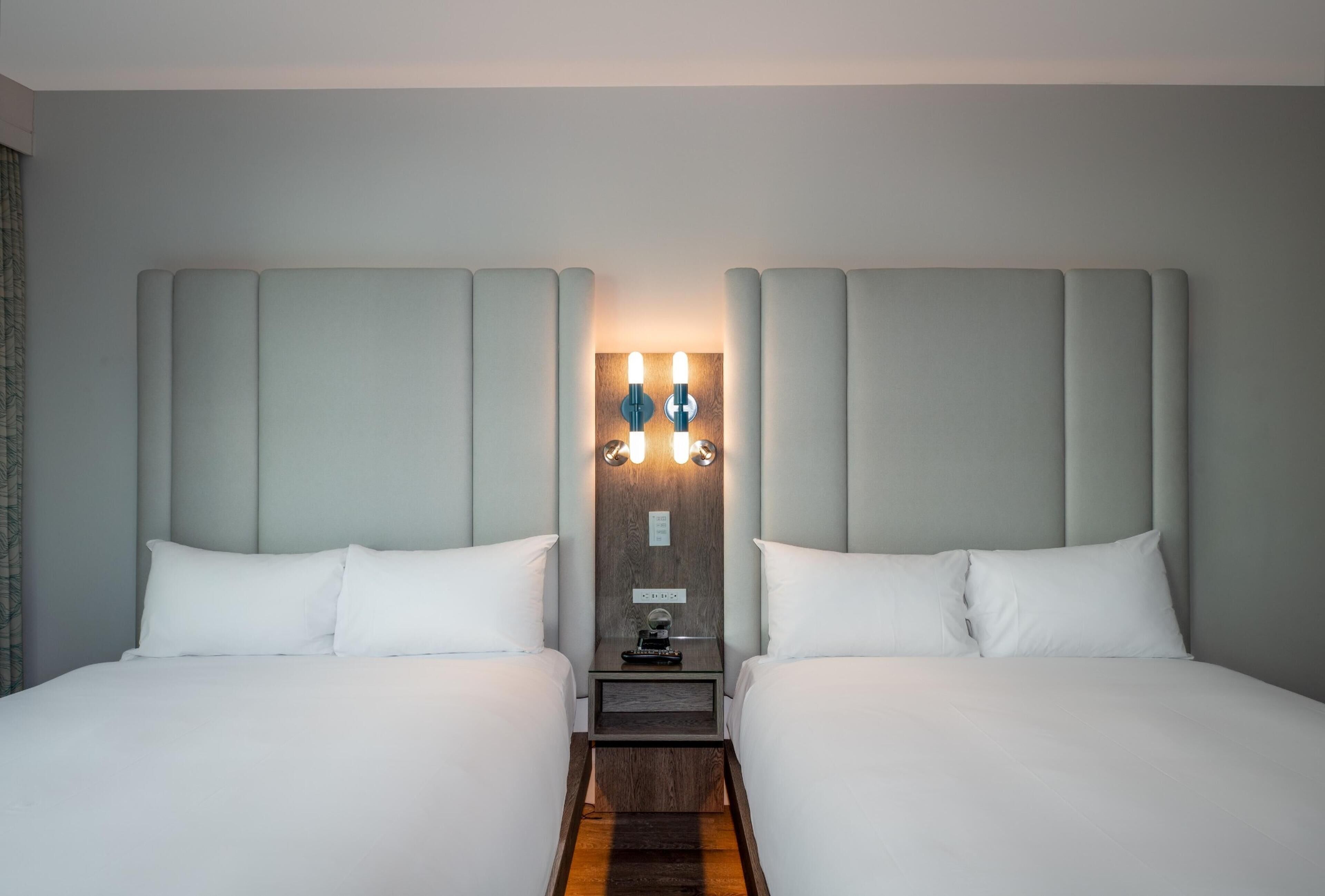 A hotel room with two beds, a nightstand with a lamp, and a wooden headboard.