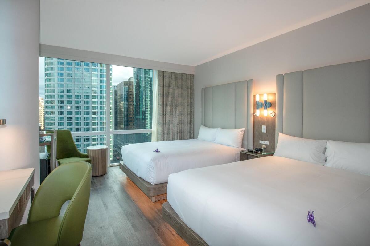 A hotel room with two beds, a desk, a chair, a round table, and a large window with a city view.
