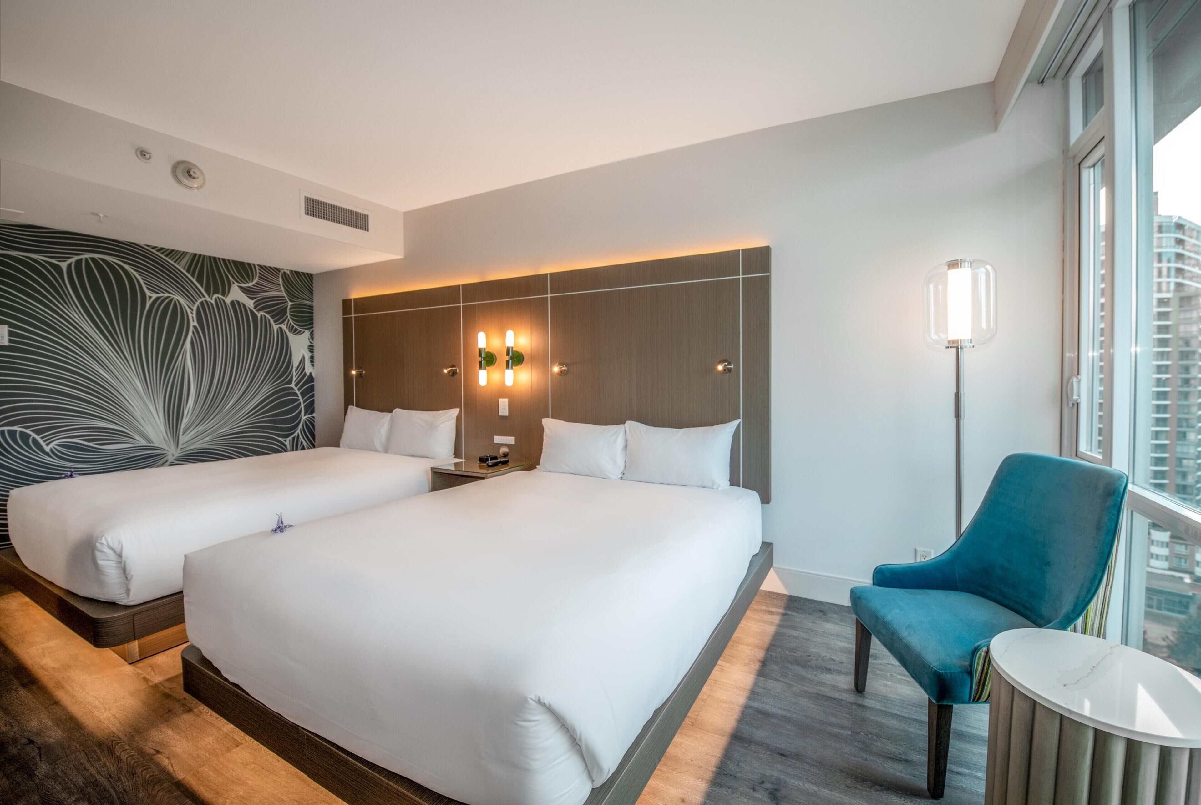 A hotel room with two beds, a blue armchair, a round table, a wall with a leaf pattern, and a city view.