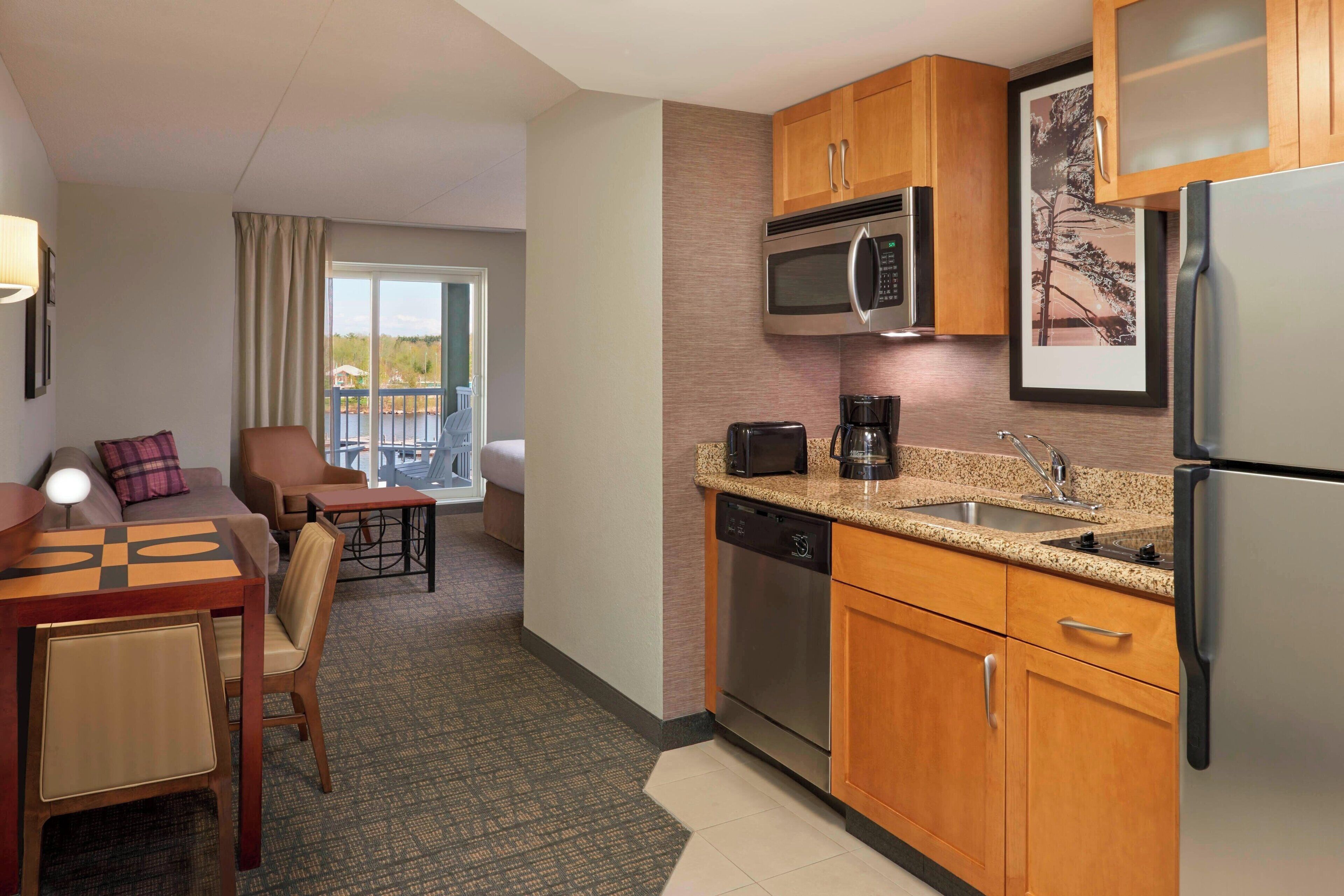 A compact hotel kitchen with wooden cabinets, a stainless steel refrigerator, a microwave, a coffee maker, and a dishwasher. There is a dining area with a table and chairs, and a view of the outdoors through a window.
