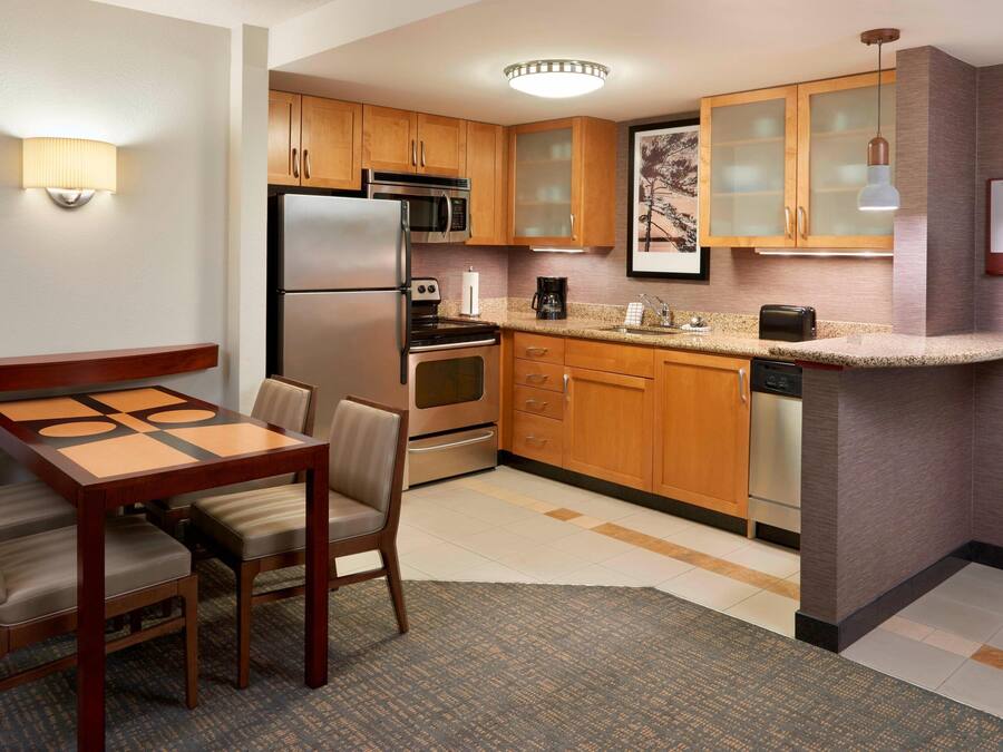 A compact kitchen with wooden cabinets, a refrigerator, microwave, oven, and a coffee maker. There is a dining table with chairs and a small rug.