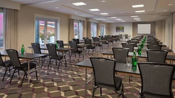 Sala de conferências com várias mesas e cadeiras, janelas amplas e tela de projeção.