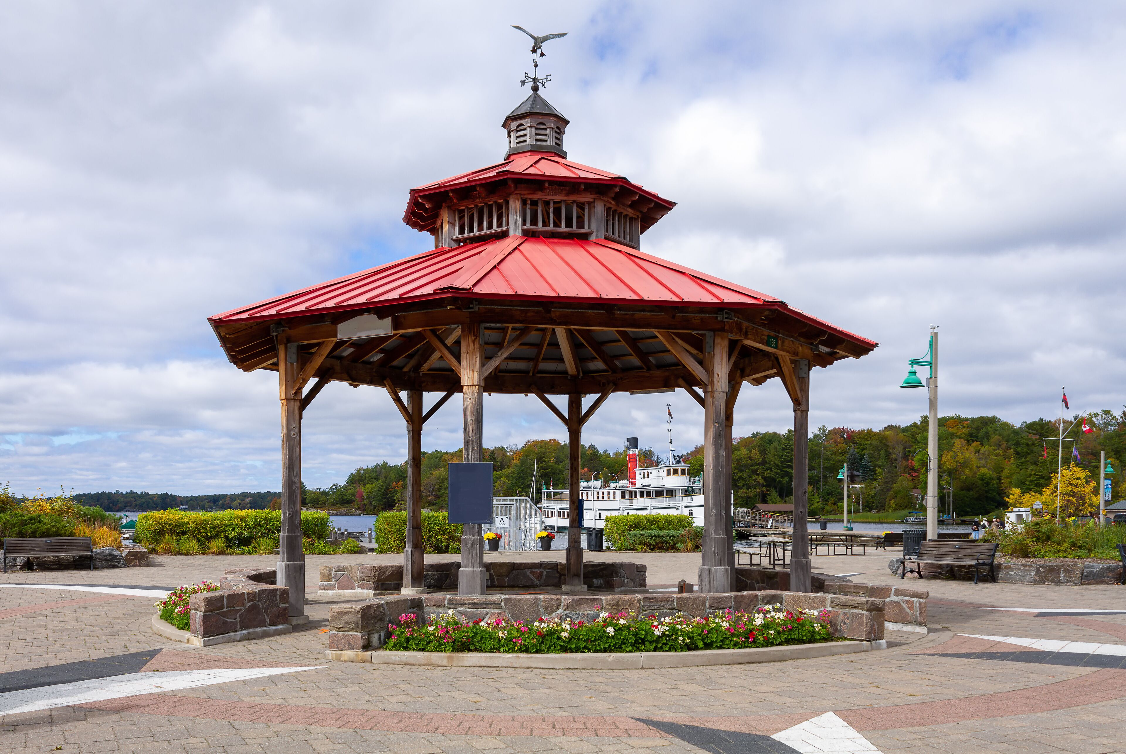 Un kiosque avec un toit rouge et une girouette, entouré de jardinières et de bancs en pierre, avec une rivière et des bateaux en arrière-plan.